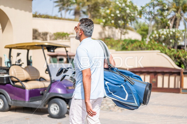 زاوية من الخلف لرجل عربي خليجي سعودي يرتدي الزي الرياضي، يحمل حقيبة الجولف، ممارسة لعبة الغولف، أنشطة رياضية بالسعودية، لعبة الجولف، رياضات الألعاب الأولمبية الصيفية، أنشطة ترفيهية، نادي الجولف، سيارة كهربائية مختصة بنقل الركاب