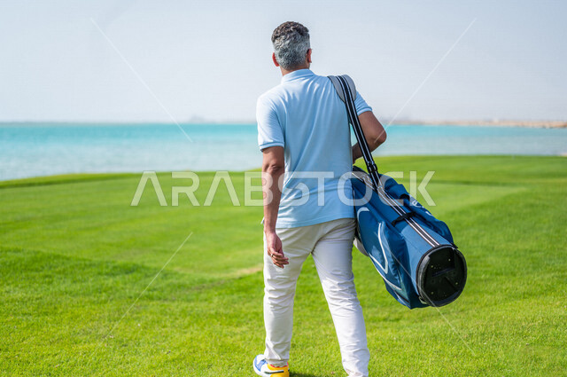 زاوية من الخلف لرجل عربي خليجي سعودي يرتدي الزي الرياضي، يحمل حقيبة الجولف، ممارسة لعبة الغولف على مساحات واسعة خضراء، أنشطة رياضية بالسعودية، لعبة الجولف، رياضات الألعاب الأولمبية الصيفية، أنشطة ترفيهية، ملعب الجولف