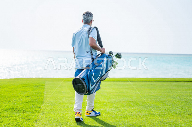 زاوية من الخلف لرجل عربي خليجي سعودي يرتدي الزي الرياضي، يحمل حقيبة الجولف، ممارسة لعبة الغولف على مساحات واسعة خضراء، أنشطة رياضية بالسعودية، لعبة الجولف، رياضات الألعاب الأولمبية الصيفية، أنشطة ترفيهية، ملعب الجولف