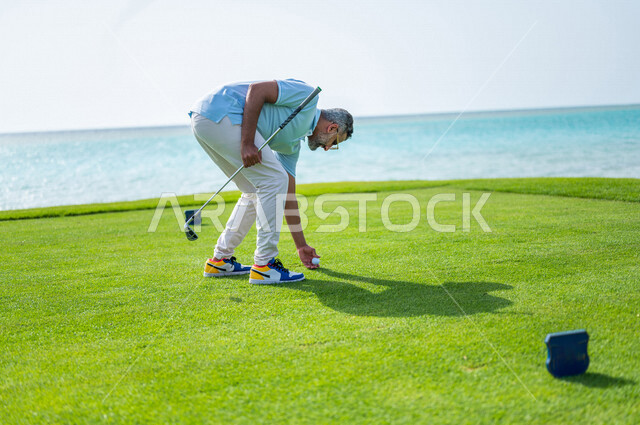 رجل عربي خليجي سعودي يرتدي الزي الرياضي، يقوم بتثبيت كرة الجولف، ممارسة لعبة الغولف على مساحات واسعة خضراء، أنشطة رياضية بالسعودية، لعبة الجولف، رياضات الألعاب الأولمبية الصيفية، أنشطة ترفيهية، ملعب الجولف، الإستمتاع باللعب