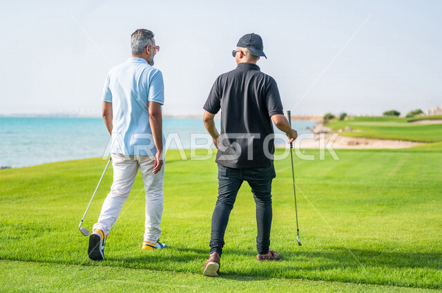 زاوية من الخلف لرجلان عربيان خليجيان سعوديان يرتديان الزي الرياضي، ممارسة لعبة الغولف على مساحات واسعة خضراء، أنشطة رياضية بالسعودية، لعبة الجولف، رياضات الألعاب الأولمبية الصيفية، أنشطة ترفيهية، ملعب الجولف