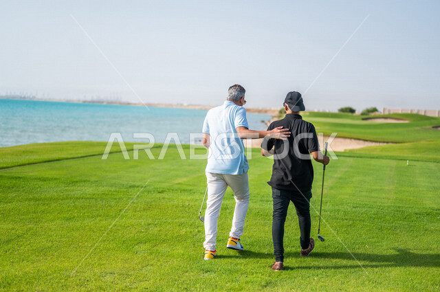 زاوية من الخلف لرجلان عربيان خليجيان سعوديان يرتديان الزي الرياضي، ممارسة لعبة الغولف على مساحات واسعة خضراء، أنشطة رياضية بالسعودية، لعبة الجولف، رياضات الألعاب الأولمبية الصيفية، أنشطة ترفيهية، ملعب الجولف
