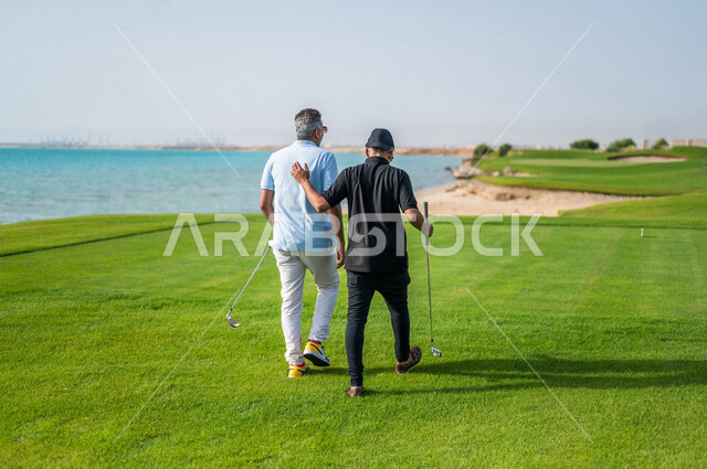 زاوية من الخلف لرجلان عربيان خليجيان سعوديان يرتديان الزي الرياضي، ممارسة لعبة الغولف على مساحات واسعة خضراء، أنشطة رياضية بالسعودية، لعبة الجولف، رياضات الألعاب الأولمبية الصيفية، أنشطة ترفيهية، ملعب الجولف