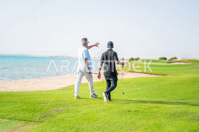 زاوية من الخلف لرجلان عربيان خليجيان سعوديان يرتديان الزي الرياضي، ممارسة لعبة الغولف على مساحات واسعة خضراء، أنشطة رياضية بالسعودية، لعبة الجولف، رياضات الألعاب الأولمبية الصيفية، أنشطة ترفيهية، ملعب الجولف