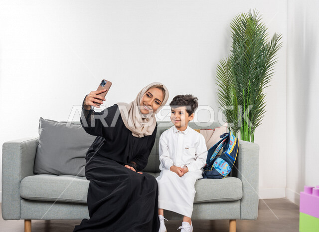 A veiled Saudi Gulf Arab mother with her son, wearing a school uniform, taking a selfie using a mobile phone, taking souvenir photos, a group photo, documenting happy moments, happy facial gestures, school backpack, new school year, back to school