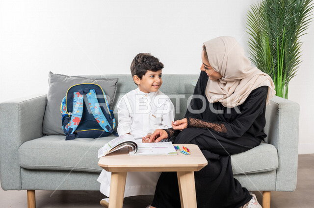 أم عربية خليجية سعودية محجبة تساعد طفلها في التعلم  بالمنزل، ايماءات وجه سعيدة، الانشطة التعليمية، تنمية قدرات الطفل، تشجيع وتطوير الأطفال، مساعدة الطفل بالواجبات المدرسية المنزلية، حقيبة الظهر المدرسية، الاهتمام ودعم الأبناء