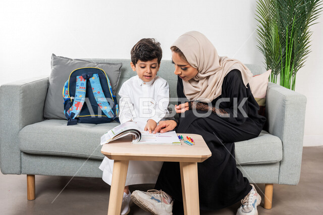 A veiled Saudi Gulf Arab mother helps her child learn at home, happy facial gestures, educational activities, developing child abilities, encouraging and developing children, helping the child with homework, school backpack, attention and support for chil