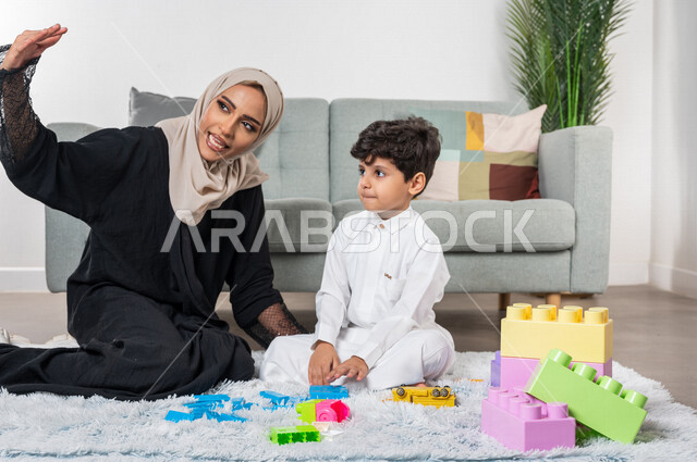 أم عربية خليجية سعودية محجبة تلعب مع طفلها، ممارسة الانشطة الترفيهية، ام وابنها يرفعون ايديهم الى اعلى بايماءات وجه سعيدة، مفهوم الأمومة والطفولة، أنشطة ترفيهية للطفل، رعاية الاطفال، تطوير مهارات الطفل، الاهتمام ودعم الأبناء