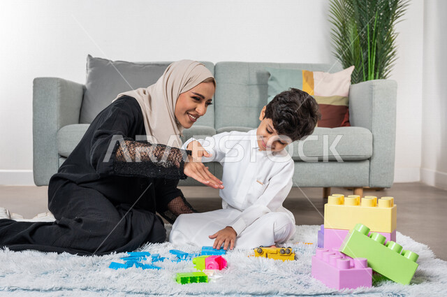 أم عربية خليجية سعودية محجبة تلعب مع طفلها، ممارسة الانشطة الترفيهية، ام وابنها يرفعون ايديهم الى اعلى بايماءات وجه سعيدة، مفهوم الأمومة والطفولة، أنشطة ترفيهية للطفل، رعاية الاطفال، تطوير مهارات الطفل، الاهتمام ودعم الأبناء