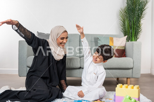أم عربية خليجية سعودية محجبة تلعب مع طفلها، ممارسة الانشطة الترفيهية، ام وابنها يرفعون ايديهم الى اعلى بايماءات وجه سعيدة، مفهوم الأمومة والطفولة، أنشطة ترفيهية للطفل، رعاية الاطفال، تطوير مهارات الطفل، الاهتمام ودعم الأبناء