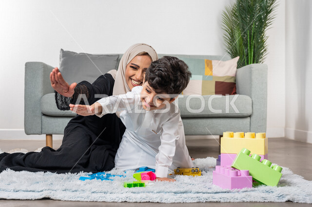 أم عربية خليجية سعودية محجبة تلعب مع طفلها، ممارسة الانشطة الترفيهية، ام وابنها يرفعون ايديهم الى اعلى بايماءات وجه سعيدة، مفهوم الأمومة والطفولة، أنشطة ترفيهية للطفل، رعاية الاطفال، تطوير مهارات الطفل، الاهتمام ودعم الأبناء