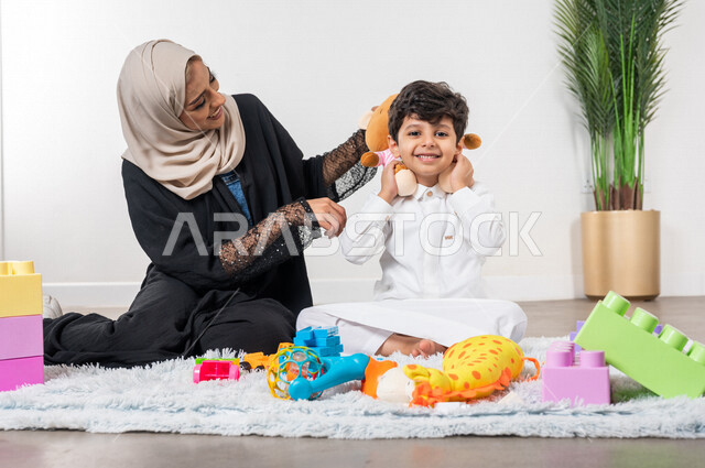 أم عربية خليجية سعودية محجبة تلعب مع طفلها، ممارسة الانشطة الترفيهية، ايماءات وجه تدل على السعادة، مفهوم الأمومة والطفولة، رعاية الاطفال، انشطة الاطفال الممتعة في المنزل، مشاركة الابناء الانشطة الترفيهية، أجواء عائلية سعيدة