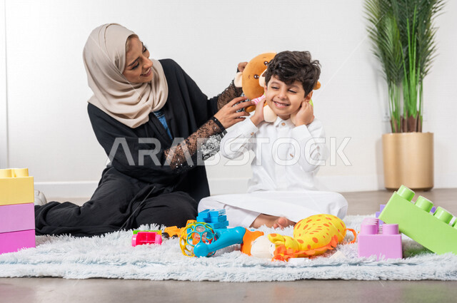 أم عربية خليجية سعودية محجبة تلعب مع طفلها، ممارسة الانشطة الترفيهية، ايماءات وجه تدل على السعادة، مفهوم الأمومة والطفولة، رعاية الاطفال، انشطة الاطفال الممتعة في المنزل، مشاركة الابناء الانشطة الترفيهية، أجواء عائلية سعيدة