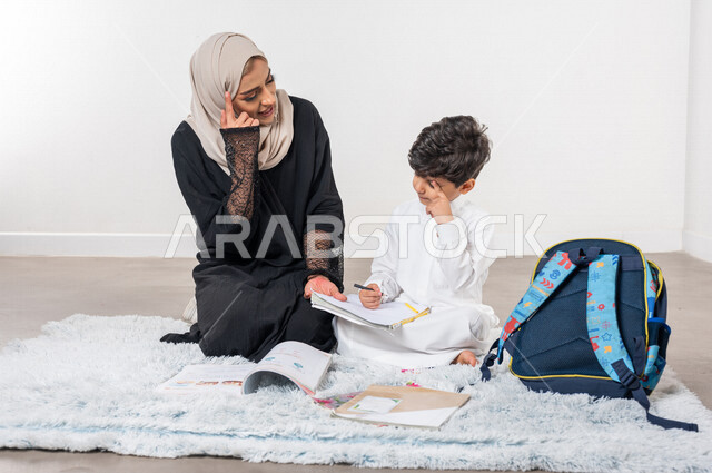 ام عربية خليجية سعودية تساعد طفلها في التعلم  بالمنزل، الانشطة التعليمية، تنمية قدرات الطفل، تشجيع وتطوير الأطفال، مساعدة الطفل بالواجبات المدرسية المنزلية، حقيبة الظهر المدرسية، ايماءات وجه ويدين تدل على التفكير،  الاهتمام و دعم  الأبناء