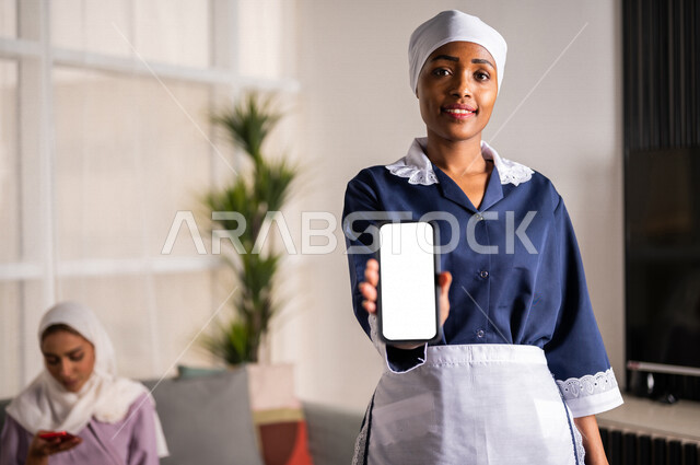 عاملة نظافة ترتدي المئزر، تحمل بيدها الهاتف المحمول، تعرض شاشة بيضاء فارغة، عرض تطبيق لخدمات عاملات التنظيف عبر الجوال، تسويق إعلانات للعمالة المنزلية، خلفية لسيدة منزل عربية سعودية تستعين بخادمة منزلية أجنبية، خدمة التنظيف المنزلي