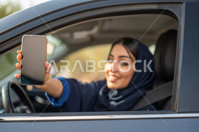 صورة مقربة لامرأه عربية خليجية سعودية محجبة تقود السيارة وتمسك بيدها الهاتف المحمول، حرية القيادة للمرأة السعودية، خدمة توصيل الركاب، خدمات النقل الامنة، ايماءات وجه مبتسمة، شاشة سوداء فارغة، عرض خدمات وتطبيقات الهاتف المحمول، التسوق الإلكتروني، قيادة الم