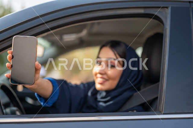 صورة مقربة لامرأه عربية خليجية سعودية محجبة تقود السيارة وتمسك بيدها الهاتف المحمول، حرية القيادة للمرأة السعودية، خدمة توصيل الركاب، خدمات النقل الامنة، ايماءات وجه مبتسمة، شاشة سوداء فارغة، عرض خدمات وتطبيقات الهاتف المحمول، التسوق الإلكتروني، قيادة الم