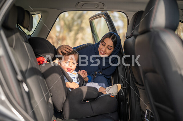صورة مقربة لامرأه عربية خليجية سعودية محجبة تقوم بوضع طفلها في السيارة، مقعد سيارة للأطفال، ايماءات وجه تدل على السعادة، الاستعداد للخروج بالسيارة، مفهوم الأمومة والطفولة، حنان الأمهات، رعاية الاطفال، خدمات النقل الامنة