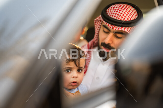 صورة مقربة لرجل عربي خليجي سعودي يجلس داخل السيارة ويلاعب طفله بسعادة، خدمة توصيل الركاب، خدمات النقل الامنة، ايماءات وجه ويدين تدل على السعادة، سيارة الاجرة، قيادة السيارات