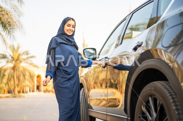 امرأه عربية خليجية سعودية محجبة تقف بجانب سيارتها، الاستعداد لركوب السيارة، خدمة توصيل الركاب، خدمات النقل الامنة، ايماءات وجه مبتسمة، صورة الجسم بالطول الكامل، الوقوف بثقة، حرية القيادة للمرأة السعودية
