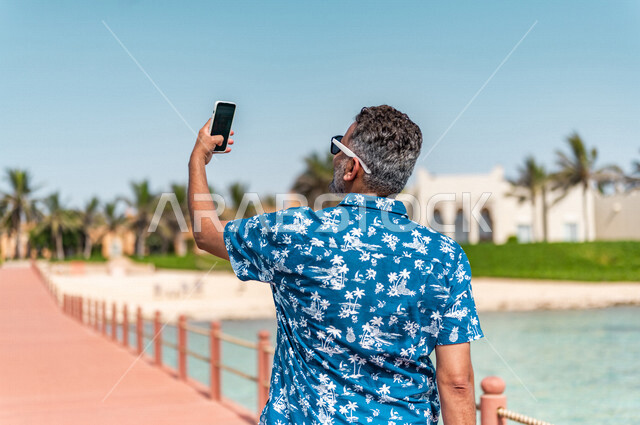صوره من الخلف لرجل عربي سعودي خليجي في رحله صيفيه ممتعه, إلتقاط صور تذكاريه عبر الهاتف المحمول, أنشطه ترفيهيه,التجول على شاطيء بحر جده, الإسترخاء والإستجمام, عطلات سياحيه, منتجع سياحي, ارتداء الملابس الكاجوال الصيفيه, السياحه في السعوديه