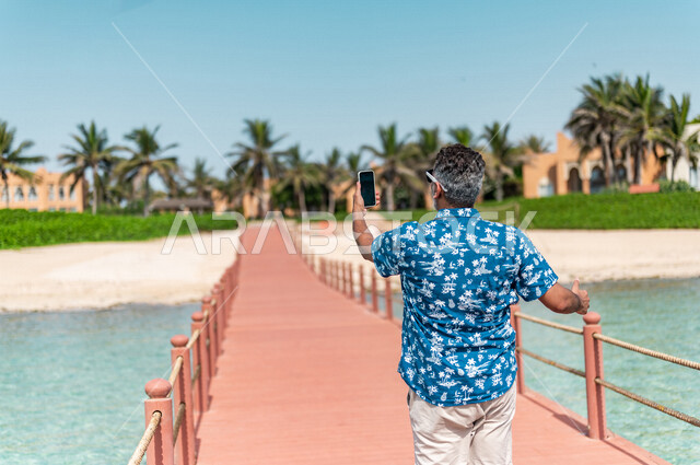 صوره من الخلف لرجل عربي سعودي خليجي في رحله صيفيه ممتعه, إلتقاط صور تذكاريه عبر الهاتف المحمول, أنشطه ترفيهيه,التجول على شاطيء بحر جده, الإسترخاء والإستجمام, عطلات سياحيه, منتجع سياحي, ارتداء الملابس الكاجوال الصيفيه, السياحه في السعوديه