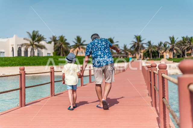A picture from the back of a Saudi Arabian Gulf man with his son on a fun summer trip, summer vacation, family entertainment activities, a tour on the beach of the Jeddah sea, taking care of children, tourist holidays, tourist resort, wearing summer cloth