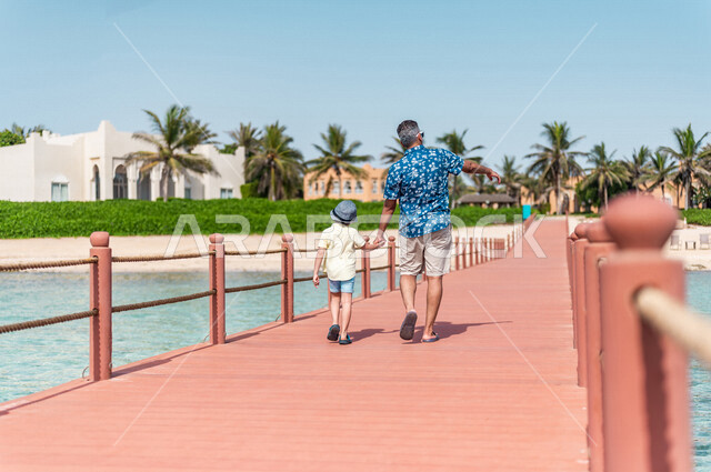 صوره من الخلف لرجل عربي سعودي خليجي مع إبنه في رحله صيفيه ممتعه, عطله صيفيه, أنشطه ترفيهيه عائليه, جوله على شاطيء بحر جده, الإهتمام بالأبناء, عطلات سياحيه, منتجع سياحي, ارتداء الملابس الصيفيه