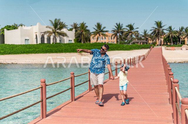 A Saudi Arabian Gulf man with his son on a fun summer trip, summer vacation, family entertainment activities, a tour on the beach of the Jeddah sea, taking care of children, tourist holidays, tourist resort, wearing summer clothes