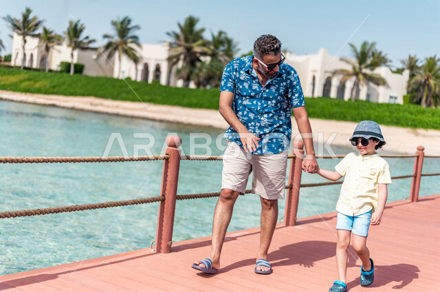 رجل عربي سعودي خليجي مع إبنه في رحله صيفيه ممتعه, عطله صيفيه, أنشطه ترفيهيه عائليه, جوله على شاطيء بحر جده, الإهتمام بالأبناء, عطلات سياحيه, منتجع سياحي, ارتداء الملابس الصيفيه