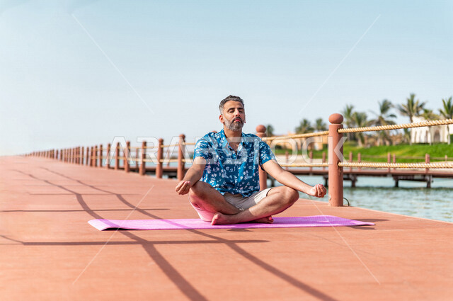 رجل عربي خليجي سعودي في رحله صيفيه ممتعه, ممارسة رياضة اليوغا على شاطئ بحر جدة، الإسترخاء والإستجمام, الرياضه الصباحيه, منتجع سياحي, عطلات سياحيه, ارتداء الملابس الكاجوال الصيفيه, السياحه في السعوديه, إجازه صيفيه