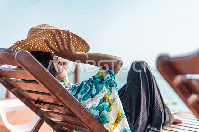 صوره من الخلف لإمراه عربيه سعوديه خليجيه على كرسي الإسترخاء على شاطيء البحر, الإستجمام والإسترخاء, منتجع سياحي, العطله الصيفيه، سياحة وترفيه، إرتداء الملابس الكاجوال الصيفيه, الإجازه الصيفيه, السياحه في السعوديه
