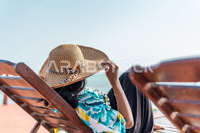 صوره من الخلف لإمراه عربيه سعوديه خليجيه على كرسي الإسترخاء على شاطيء البحر, الإستجمام والإسترخاء, منتجع سياحي, العطله الصيفيه، سياحة وترفيه، إرتداء الملابس الكاجوال الصيفيه, الإجازه الصيفيه, السياحه في السعوديه