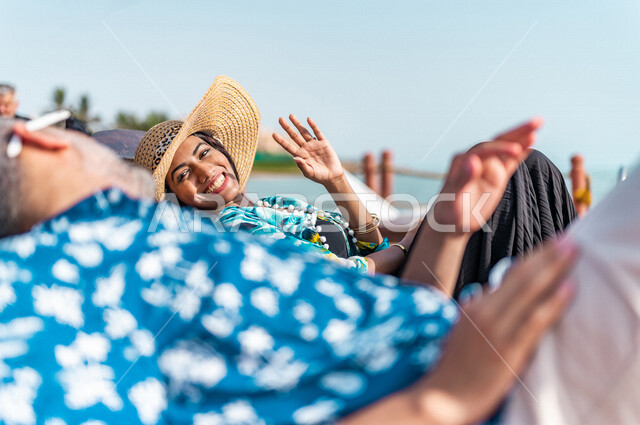 زوجان عربيان سعوديان خليجيان يجلسان على كرسي الإسترخاء على شاطيء البحر, عطلة متزوجين في شهر العسل, وجهات سياحيه لشهر العسل, منتجع سياحي، سياحة وإستجمام، أناقه شاطئيه صيفيه ملونه, إجازه صيفيه, أجواء رومانسيه