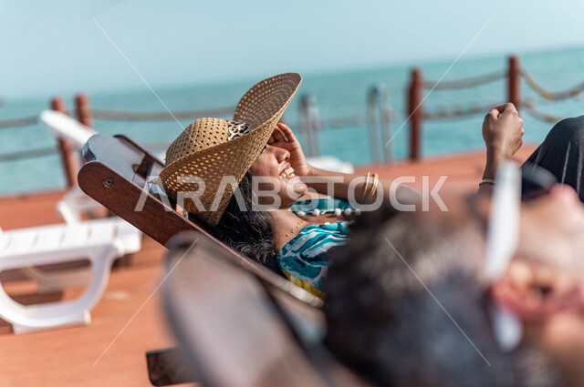زوجان عربيان سعوديان خليجيان يجلسان على كرسي الإسترخاء على شاطيء البحر, عطلة متزوجين في شهر العسل, وجهات سياحيه لشهر العسل, منتجع سياحي، سياحة وإستجمام، أناقه شاطئيه صيفيه ملونه, إجازه صيفيه, أجواء رومانسيه