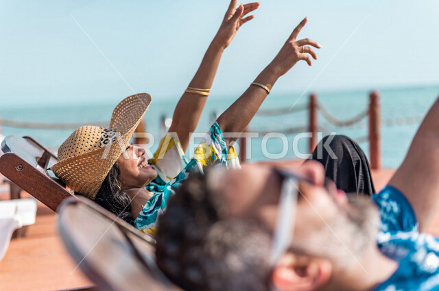 زوجان عربيان سعوديان خليجيان يجلسان على كرسي الإسترخاء على شاطيء البحر, عطلة متزوجين في شهر العسل, وجهات سياحيه لشهر العسل, منتجع سياحي، سياحة وإستجمام، أناقه شاطئيه صيفيه ملونه, إجازه صيفيه, أجواء رومانسيه
