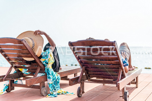 صوره من الخلف لزوجان عربيان سعوديان خليجيان يجلسان على كرسي الإسترخاء على شاطيء البحر, عطلة متزوجين في شهر العسل, وجهات سياحيه لشهر العسل, منتجع سياحي، سياحة وإستجمام، أناقه شاطئيه صيفيه ملونه, إجازه صيفيه, أجواء رومانسيه