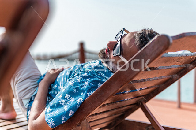 صوره من الخلف لرجل عربي سعودي خليجي على كرسي الإسترخاء على شاطيء البحر, الإستجمام والإسترخاء, منتجع سياحي, العطله الصيفيه، سياحة وترفيه، إرتداء الملابس الكاجوال الصيفيه, الإجازه الصيفيه, الجلوس تحت اشعة الشمس