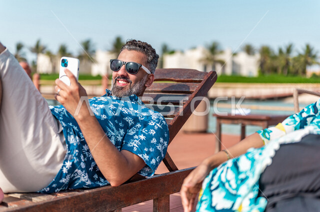 زوجان عربيان سعوديان خليجيان يجلسان على كرسي الإسترخاء على شاطيء البحر, التقاط صور تذكاريه عبر الهاتف المحمول, عطلة متزوجين في شهر العسل, وجهات سياحيه لشهر العسل, منتجع سياحي، سياحة وإستجمام، أناقه شاطئيه صيفيه ملونه