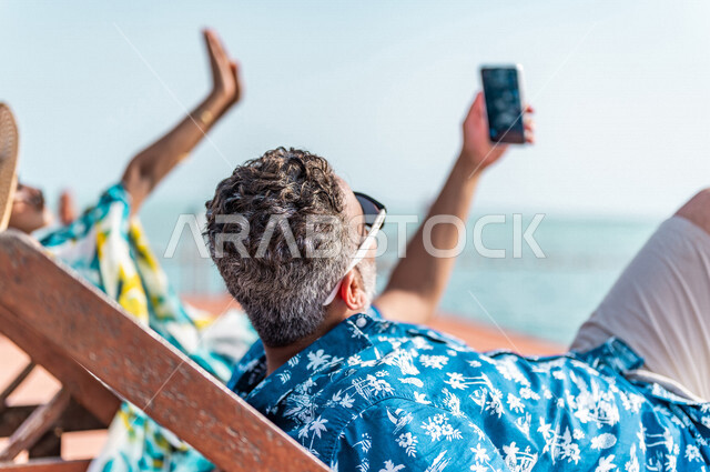 زوجان عربيان سعوديان خليجيان يجلسان على كرسي الإسترخاء على شاطيء البحر, التقاط صور تذكاريه عبر الهاتف المحمول, عطلة متزوجين في شهر العسل, وجهات سياحيه لشهر العسل, منتجع سياحي، سياحة وإستجمام، أناقه شاطئيه صيفيه ملونه