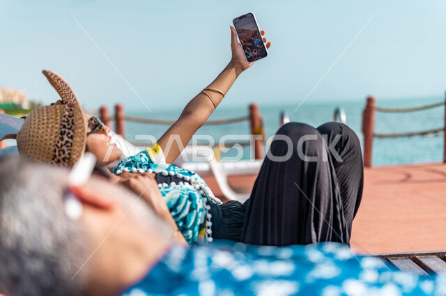 زوجان عربيان سعوديان خليجيان يجلسان على كرسي الإسترخاء على شاطيء البحر, التقاط صور تذكاريه عبر الهاتف المحمول, عطلة متزوجين في شهر العسل, وجهات سياحيه لشهر العسل, منتجع سياحي، سياحة وإستجمام، أناقه شاطئيه صيفيه ملونه
