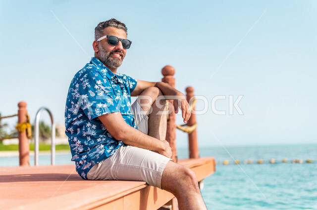 A Saudi Arabian Gulf man on a fun summer trip, summer vacation, recreational activities, sitting on the beach of the Jeddah sea, summer activities, relaxation and recreation, feeling happy, tourist holidays, tourist resort, wearing summer casual clothes, 