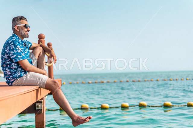 A Saudi Arabian Gulf man on a fun summer trip, summer vacation, recreational activities, sitting on the beach of the Jeddah sea, summer activities, relaxation and recreation, feeling happy, tourist holidays, tourist resort, wearing summer casual clothes, 