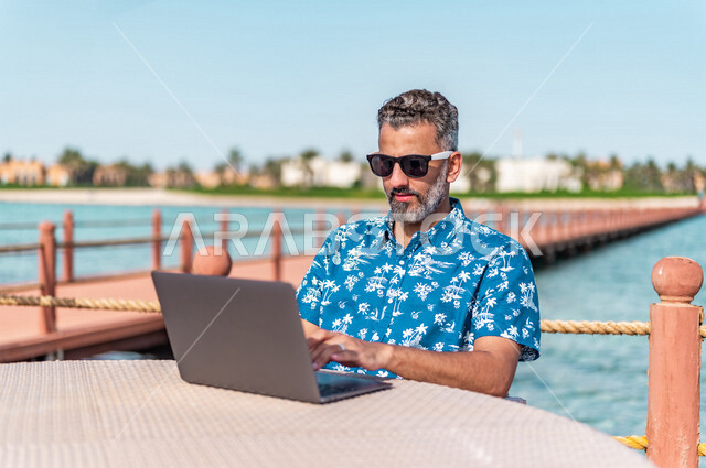 A Saudi Arabian Gulf man in Jeddah Corniche walkway, using his laptop, working during summer vacation, business trip, summer business, online shopping, making a video call over the Internet, summer vacation, wearing summer casual clothes