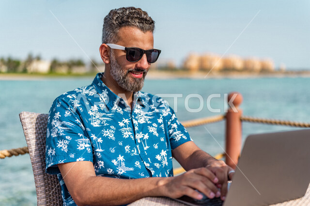 رجل عربي سعودي خليجي في ممشى كورنيش جده, إستخدام جهاز الحاسوب المحمول, العمل أثناء الإجازه الصيفيه, رحلة عمل, الأعمال الصيفيه, التسوق الإلكتروني, إجراء مكالمة فيديو عبر الانترنت, عطله صيفيه, إرتداء الملابس الكاجوال الصيفيه