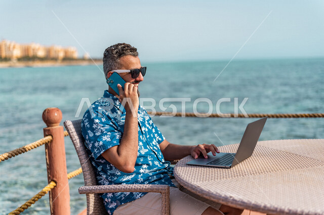 رجل عربي سعودي خليجي في ممشى كورنيش جده, إستخدام جهاز الحاسوب المحمول, إجراء مكالمه هاتفيه عبر الهاتف المحمول, العمل أثناء الإجازه الصيفيه, أعمال صيفيه, التسوق الإلكتروني, إرتداء الملابس الكاجوال الصيفيه, رحلة عمل