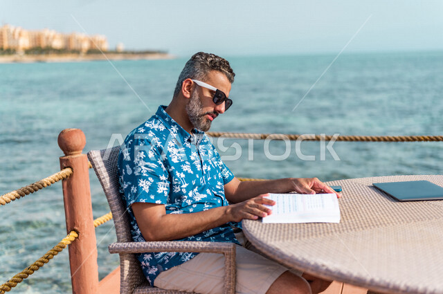 رجل عربي سعودي خليجي في ممشى كورنيش جده, الإستمتاع بالقراءه, تصفح الكتاب, الثقافه والتعليم, عطله صيفيه, إرتداء الملابس الكاجوال الصيفيه, السياحه في المملكه العربيه السعوديه