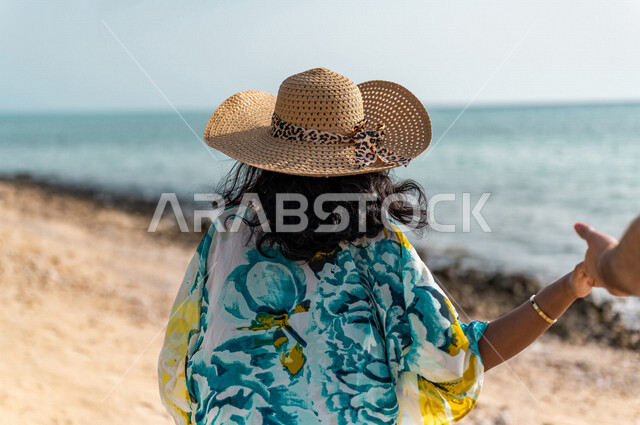 صوره من الخلف لإمراه عربيه سعوديه خليجيه تمسك بيد زوجها على شاطيء البحر, أجواء رومانسيه, عطلة متزوجين في شهر العسل, وجهات سياحيه في شهر العسل، المشي على رمال البحر، سياحة وإستجمام، أناقه شاطئيه ملونه, الإجازه الصيفيه