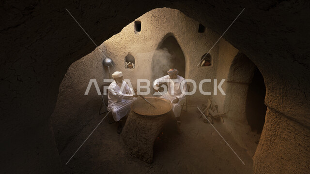 صورة لرجلان عربيان خليجيان عمانيان يرتديان الزي العماني ، صنع الحلوى العمانية ، الحلويات العمانية قديما ، المرجل النحاسي ، موقد تراثي من الطين ، مباني طينية قديمة ، شعبيات