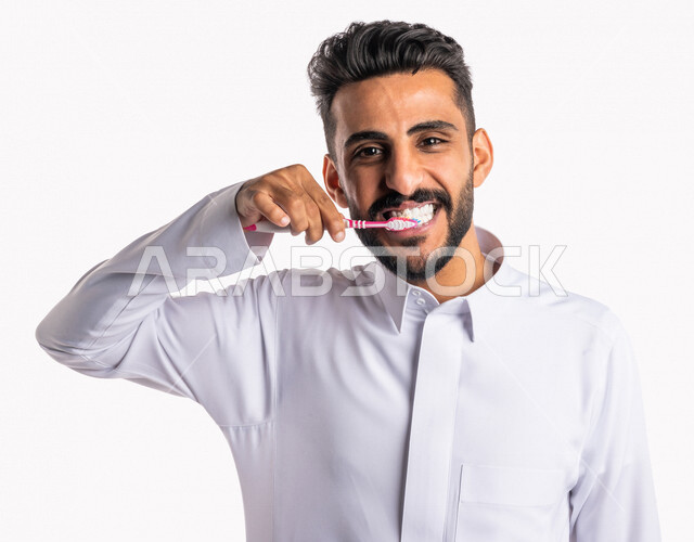 بورتريه بصورة مقربة لرجل عربي خليجي سعودي ، يقوم بتنظيف الأسنان بفرشاة الأسنان ، مفهوم العناية والإهتمام بصحة ونظافة الأسنان ، روتين نظافة الفم اليومية ، تجنب تسوس الاسنان وامراض اللثة ، خلفية بيضاء